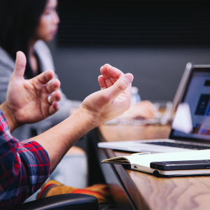Hand gestures in the foreground with an employee in the background sitting at a table having a meeting